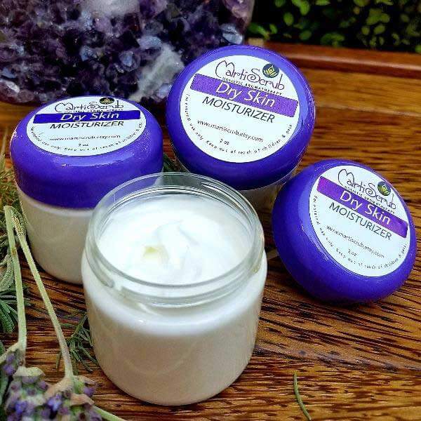 Three jars of Dry Skin Moisturizer with organic ingredients, featuring Argan oil and essential oils, displayed outdoors.